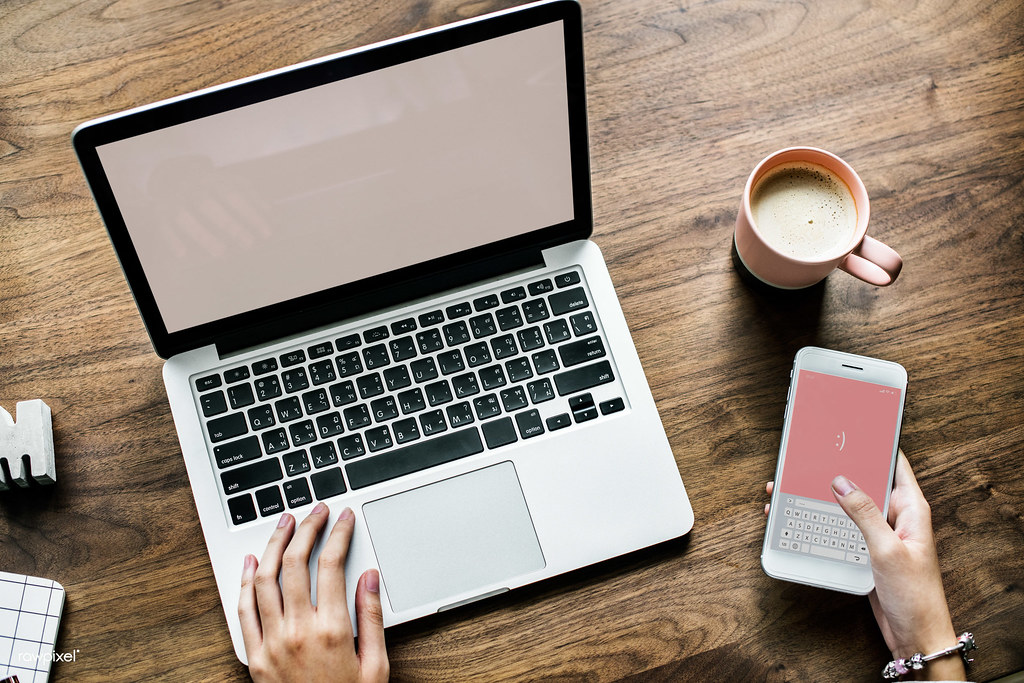 Hands on a laptop keyboard beside a smartphone and a cup of coffee on a wooden desk
